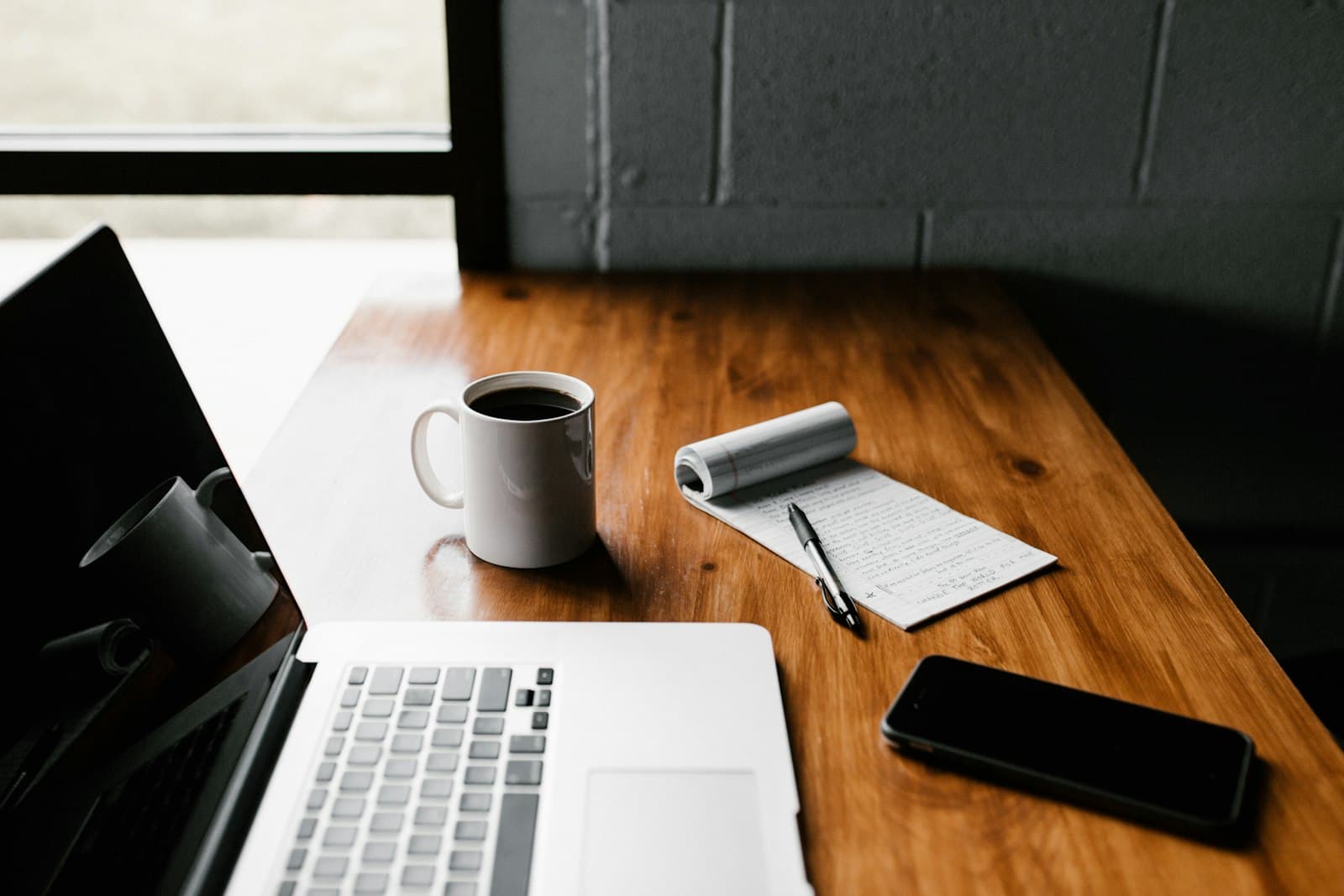A laptop, coffee mug, and notebook on a warm wooden desk by a window