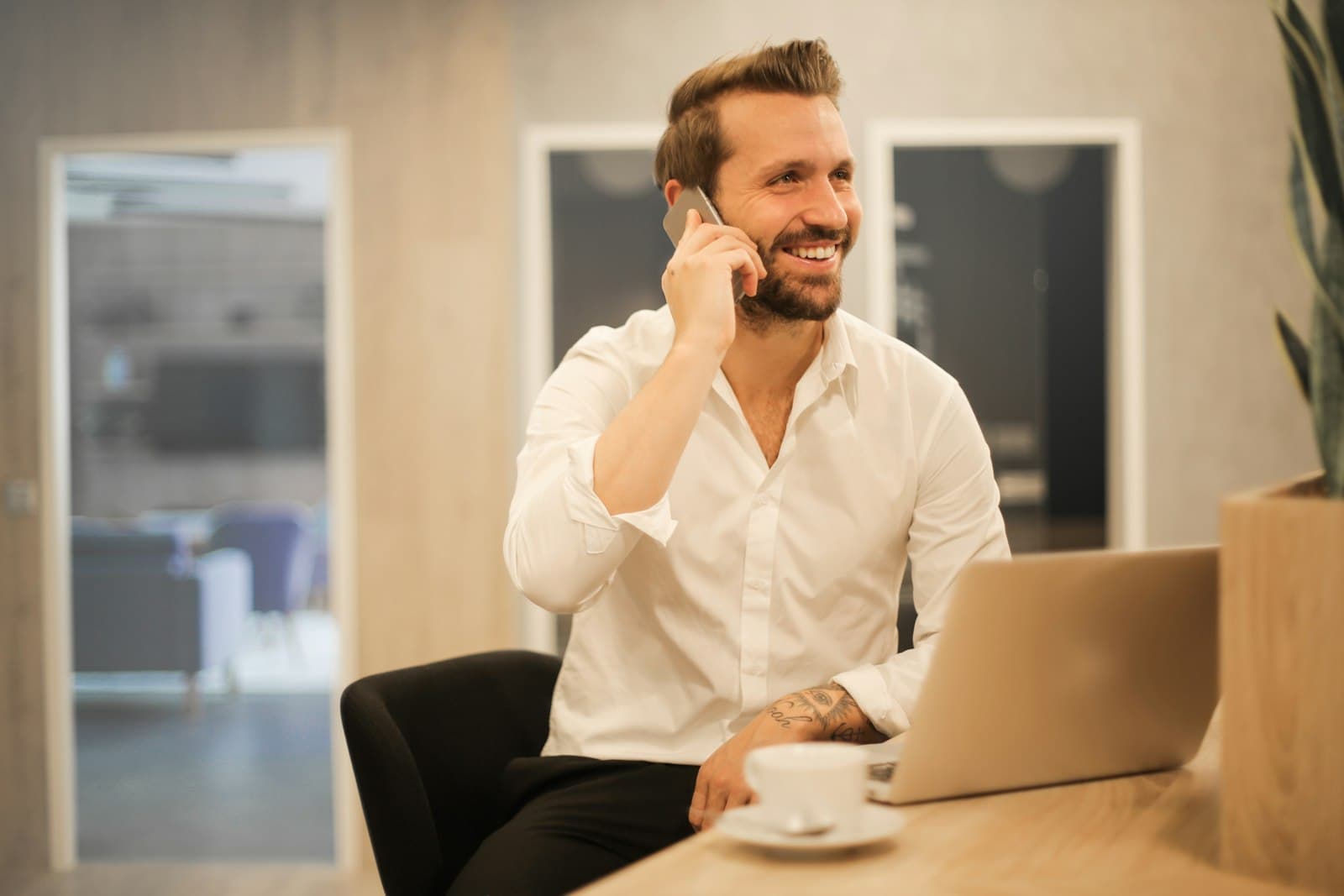 Business owner in a white shirt smiling on the phone at a desk with a laptop and coffee